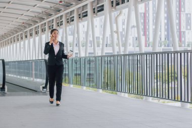 young Asian businesswoman in black suit using phone and holding cup of coffee and standing on elevated walkway station with copy space.