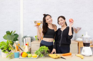 Asian active two women sibling in sportswear enjoy eating fruits healthy foods. Beautiful girl, sister holding a glass of orange juice and apples in the kitchen.