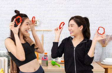 Asian active two women sibling in sportswear enjoy eating fruits healthy foods.Beautiful girl sister holding Bell Pepper, sliced like glasses. In the kitchen