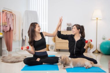 İki güzel Asyalı kız spor kıyafetleri giyer ve yatak odasında köpeklerle gülümserler. Dambılı ve tabletli kadın egzersiz paspası