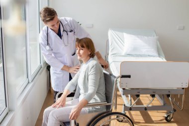 Kafkasyalı erkek doktor, tekerlekli sandalyedeki yaşlı kadın hastayı muayene ediyor..