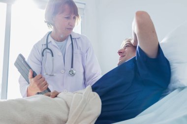 Doktor, hastanedeki hastayı muayene ediyor. Kafkasyalı kadın doktor hastane yatağında yatan erkek hastayla konuşuyor..