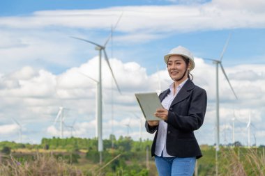 Rüzgar çiftliğinde çalışan enerji mühendisi kadın. Asyalı iş kadını elinde tablet ve panoyla yel değirmeni ortamında yenilenebilir enerji taşıyor..