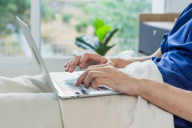 Hastane odasında dizüstü bilgisayar kullanan beyaz bir hasta. Hasta bir adam hastane yatağında yatıyor..