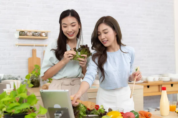 stock image Two young Asian women are using a tablet in the kitchen. Woman having fun with healthy food salad.