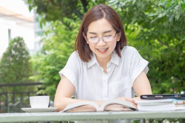 Asian middle aged woman in glasses reading a book in backyard.
