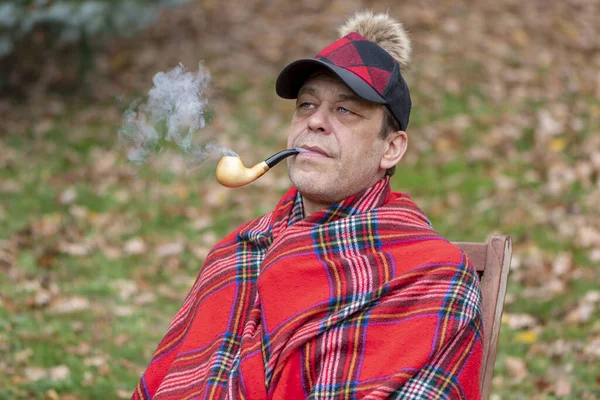 Stylish man 45-50 years old smokes a pipe, sits at a table, in a plaid and a cap with a pompom on a blurred background of nature.