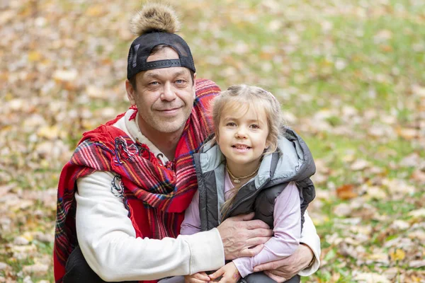 Family portrait of a stylish man 45-50 years old in a plaid hugs his daughter 4-5 years old against the background of grass and fallen leaves.