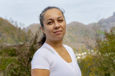 Dağların ve ormanların bulanık arka planında beyaz tişörtlü 40-45 yaşlarında bir kadının portresi. Kavram: aktif eğlence, turizm ve seyahat.