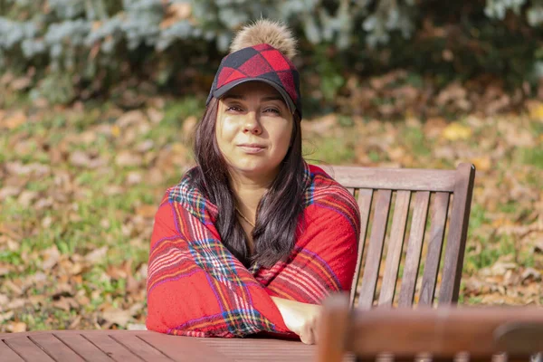 stock image Street portrait of a stylish 40-45-year-old brunette woman wrapped in a red plaid plaid, sitting in a wooden garden chair against the background of nature.