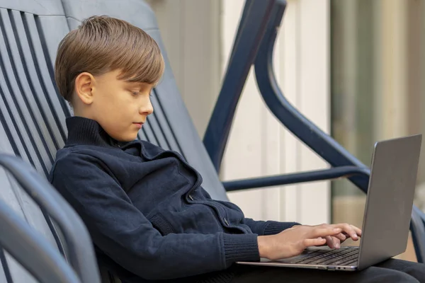 A 10-year-old boy is sitting in a plastic chair, typing on a laptop. Concept: remote learning, communication via the Internet, homework.