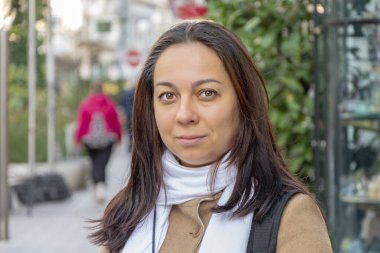 40-45 yaşlarında güzel bir esmer kadın kahverengi ceketli ve şehir yeşilliğinin bulanık arka planında beyaz bir eşarp takıyor..