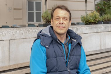 45-50 yaşlarındaki yaşlı bir adamın sokak portresi bulanık bir şehir geçmişine karşı bankta oturuyor..