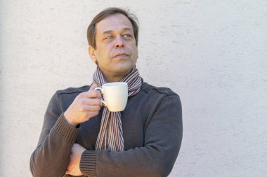 45-50 yaşlarında, eşarplı, şık, yaşlı bir adam beyaz bir fincanı elinde tutuyor..