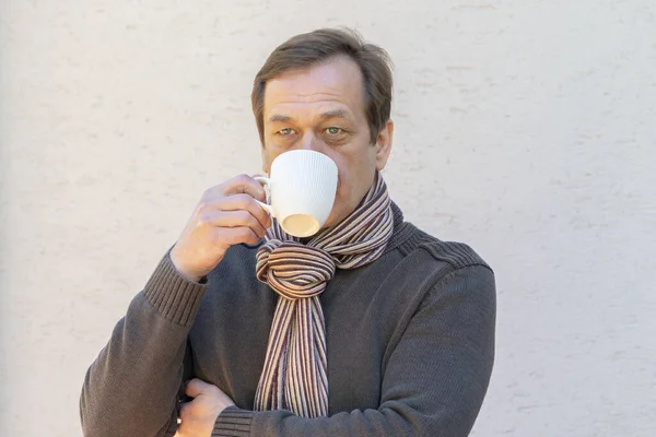 An elderly stylish man 45-50 years old in a scarf drinks from a white cup on a light background.