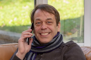 Eşarplı yaşlı bir adam akıllı telefonuyla koltukta oturmuş kameraya bakıyor..