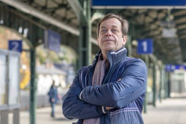 45-50 yaşındaki ceketli bir adamın portresi tren ya da tren beklerken kolları göğsünde çaprazlanmış bir tren istasyonunun platformunda duruyor..