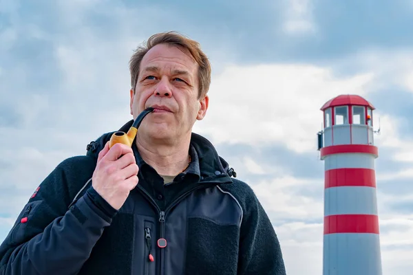 A 45-50-year-old man smokes a pipe against the background of a lighthouse and a blue sky with clouds.
