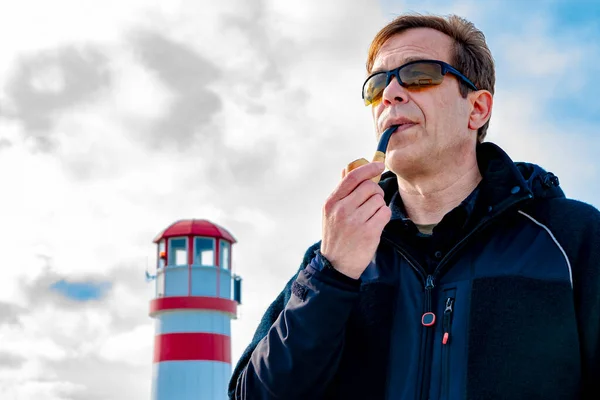 An elderly man 45-50 years old smokes a pipe against the background of a lighthouse and a blue sky with clouds, a lighthouse keeper.