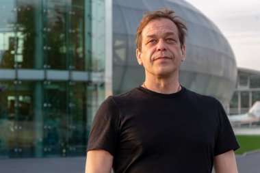 Kırk-elli yaşlarında, cam bir binanın arka planında siyah tişörtlü bir adamın sokak portresi. Belki de bir turist, gezgin, aktör, kamyon şoförü, yükleyici ya da emeklidir.,