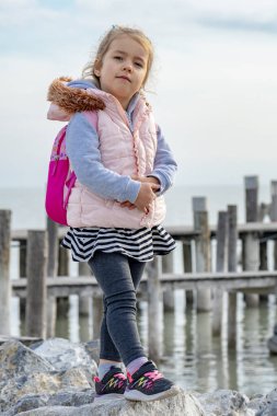 Güzel bir kız sırtında sırt çantasıyla kayaların üzerinde yürüyor, ellerini denizin ve gökyüzünün arka planına çarpıyor. ahşap köprü.
