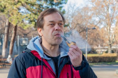 45-50 yaşlarında ceketli yaşlı bir adam bulanık bir şehir geçmişinde ahşap bir pipo içiyor..