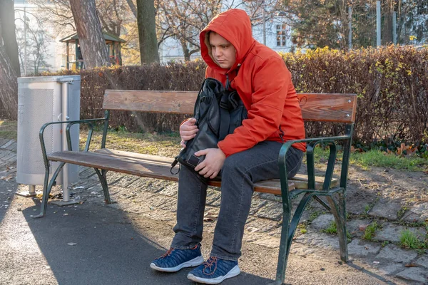 Lonely and sad guy 25-30 years old with a backpack in the park on a bench, blurred urban background. Concept: a homeless man on the street, a small unemployment benefit, life troubles.