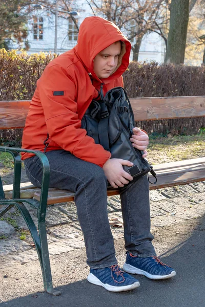 a frozen guy 25-30 years old with a backpack in the park on a bench, a blurry urban background. Concept: a homeless man on the street, a small unemployment benefit.