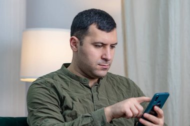 A man of 30-35 years of oriental appearance looks into a smartphone while sitting on the couch.