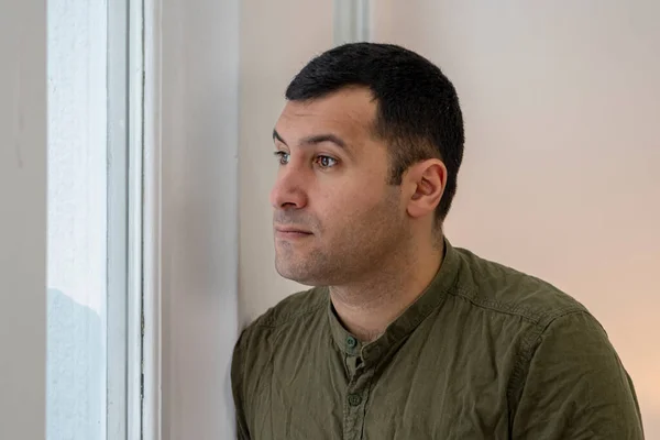 a 30-35-year-old man looks out the window on a light background.