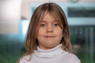 Bulanık bir arkaplanda, uzun sarı saçlı ve beyaz kazaklı 4 yaşındaki küçük bir kızın portresi..