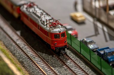 A toy electric train with a train train passes by a road with cars.