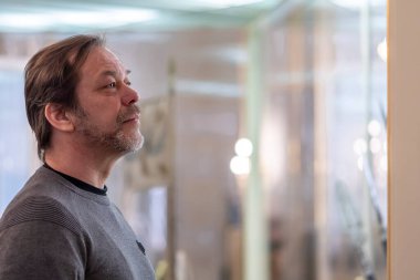 An elderly man with a beard of 45-50 years old looks seriously and attentively at something on a blurry background in the room.