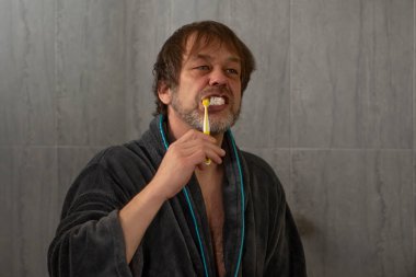 An elderly man 45-50 years old is brushing his teeth with a toothbrush on a blurry gray background in a bathrobe.