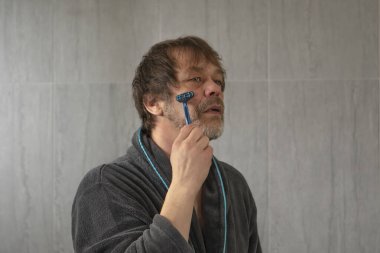 An elderly man 45-50 years old shaves off his beard on a blurry gray background in a bathrobe.