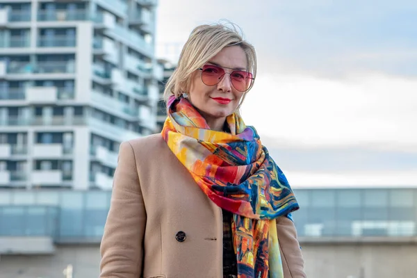 stock image Portrait of a blonde 40-45 years old in a coat on a blurry background of urban multi-storey buildings, street photography.