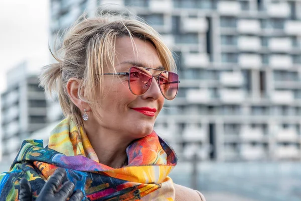 A beautiful WOMAN of 40-45 years old with glasses and a scarf on the background of urban multi-storey buildings.