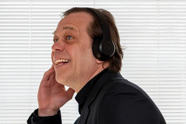 一个拿着耳机在白百叶窗背景下听音乐的老年男子 一个40 50岁的音乐爱好者的情感肖像 — 图库照片