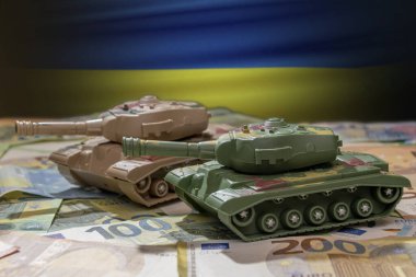 Tanklar, Ukrayna bayrağının arka planına karşı avro banknotlarıyla ilerliyor. Kavram: askeri ve mali yardım, silah maliyetleri, Rusya ile savaş, silah desteği, teçhizat transferi.