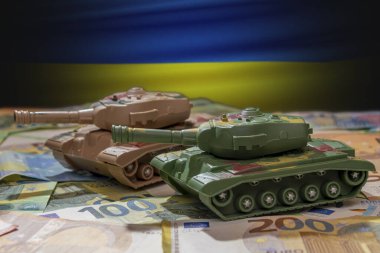 Tanklar, Ukrayna bayrağının arka planına karşı avro banknotlarıyla ilerliyor. Kavram: askeri ve mali yardım, silah maliyetleri, Rusya ile savaş, silah desteği, teçhizat transferi.