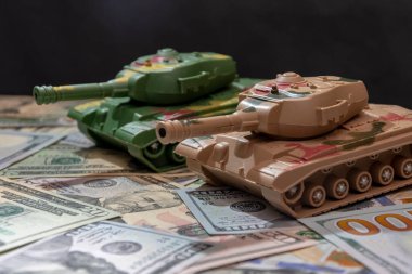 Toy military tanks, scattered american dollars, black background.  Concept: arms spending, military aid, arms and ammunition trade, money loan, war in Ukraine. clipart