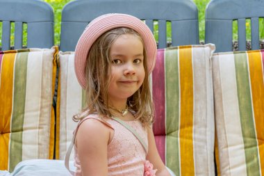 Pembe şapkalı bir kız gülümser ve renkli minderlere oturur, bir sokak portresi.