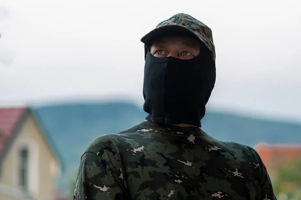 一个身穿制服和巴拉克拉瓦的士兵在模糊的山区和房屋背景上 军队动员 前线雇佣军 乌克兰战争 — 图库照片