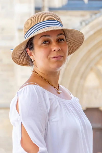 A smiling middle-aged woman wearing a straw hat against a backdrop of old European architecture