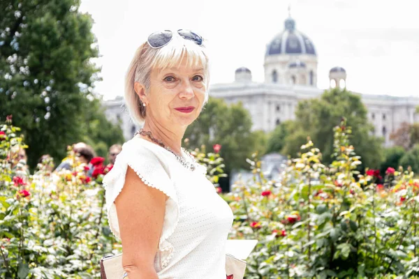 Portrait of a woman 60-65 years old in summer on a blurred background of a city garden with flowers, ancient architecture of the Austrian city of Vienna, tourism and travel.