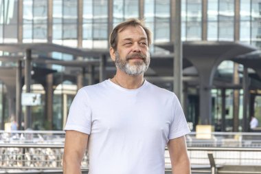 50-55 yaşlarında, cam bir binanın arka planında gri sakallı, güçlü yapılı, kaslı bir adam..