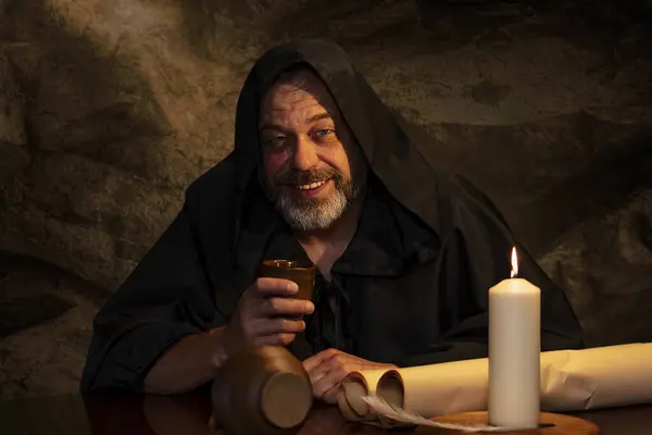 Cheerful monk in cassock drinking wine from a clay goblet sitting at a table, a jug of wine and drinking in a medieval inn. Concept: drunkenness and alcoholism, dinner in an inn.