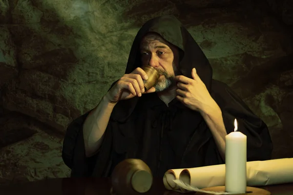 Monk in black cassock drinking wine from clay goblet sitting at table, jug of wine and drinking in medieval inn. Concept: drunkenness and alcoholism, dinner in an inn.