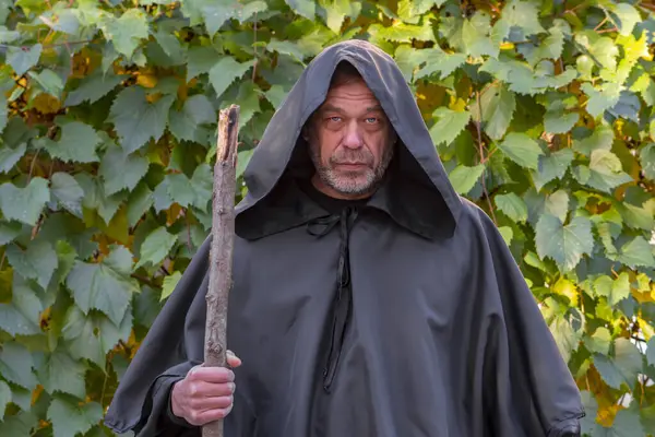 Severe Monk in a black robe with a staff on a background of grape leaves. Mysticism and magic, sorcerers and witches. Wine production in a monastery by monks.