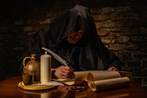 An old monk in his cell writes a message on parchment with a goose quill pen by candlelight